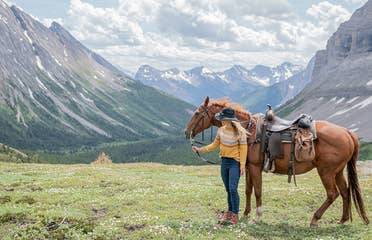 Featured Contributor, Ashlyn George, stands with a horse in front of the Canadian mountain range.