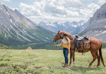 Featured Contributor, Ashlyn George, stands with a horse in front of the Canadian mountain range.