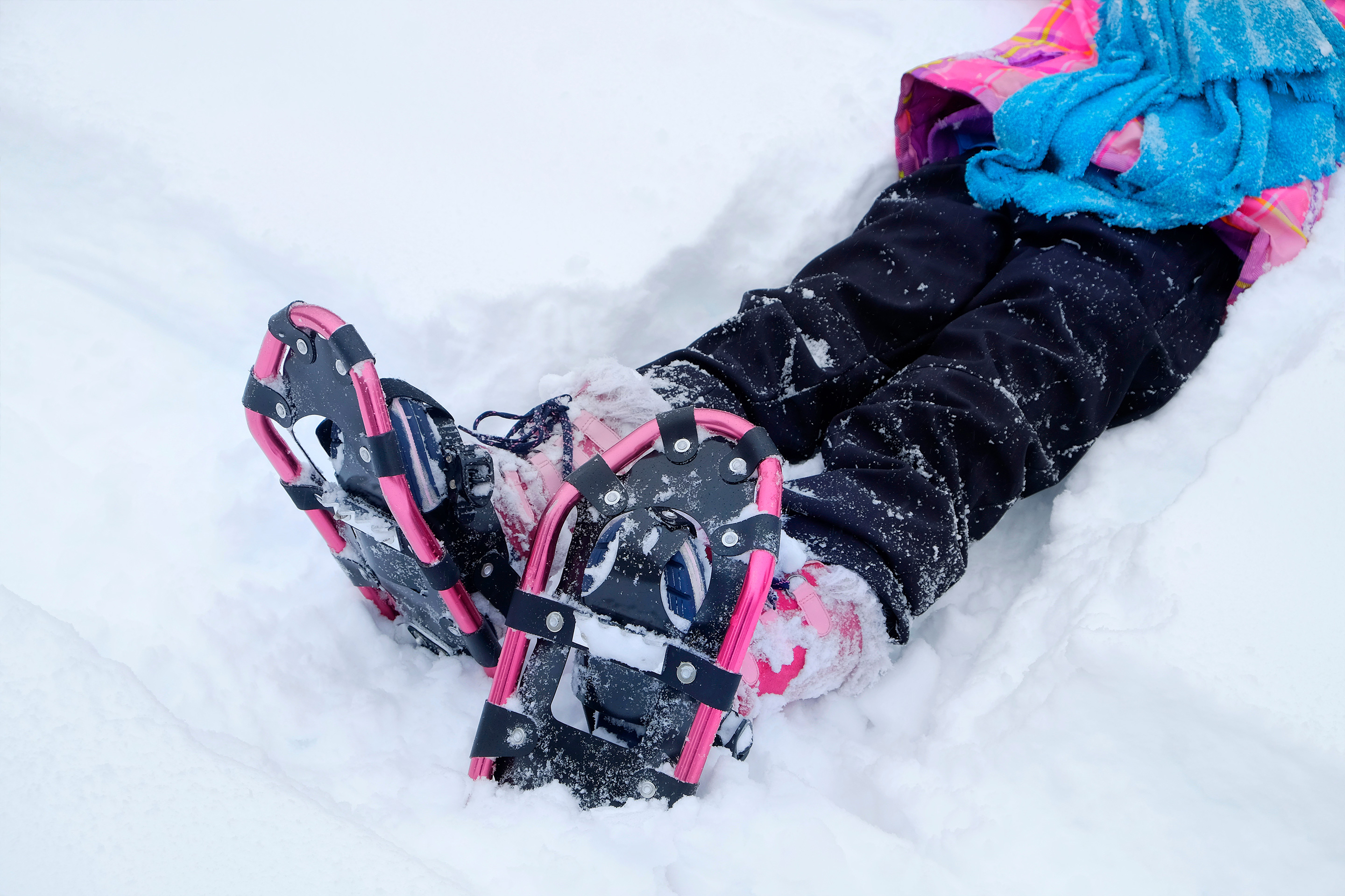 A girl sits in the snow wearing a blue and pink jacket, black snow pants and a pair of pink and black snowshoes.