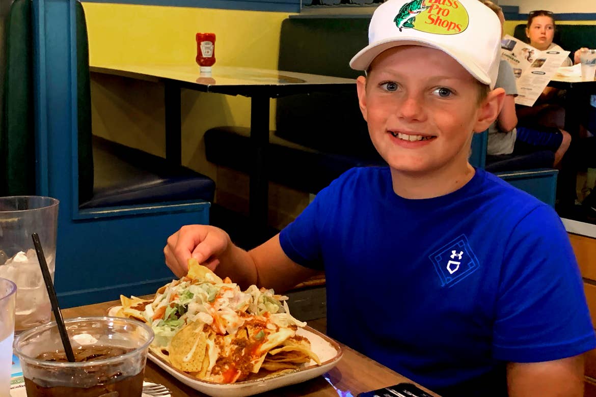A young boy in a blue t-shirt and white baseball cap sits at a dining table with a plate of nachos.