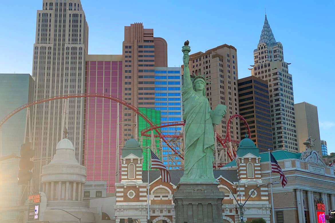 A scaled 'Statue of Liberty' and New York Skyline has red rollercoaster tracks intertwined in the air under a blue sky.