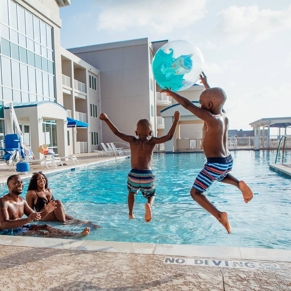 A woman, man and two young boys jump into an infinity pool near a beachfront with a beach ball.