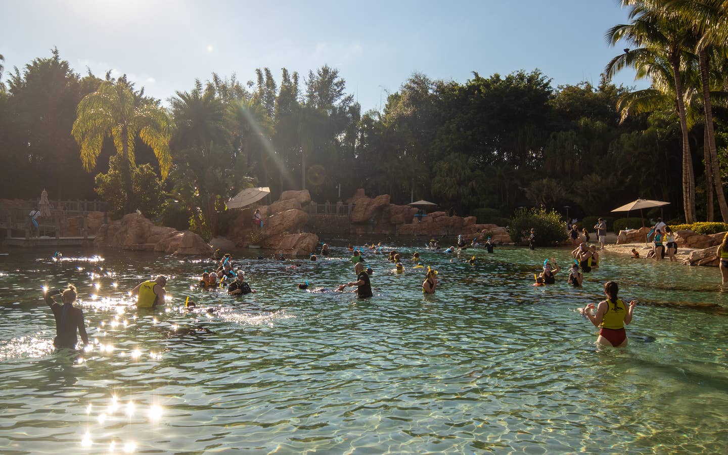The sun shines on an outdoor water area containing tens of guests in swimwear and lifejackets.