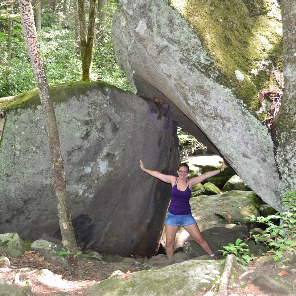 A woman wearing a purple tank top, jean shorts and glasses poses underneath a pair of wedged rocks in Gatlinburg, TN.
