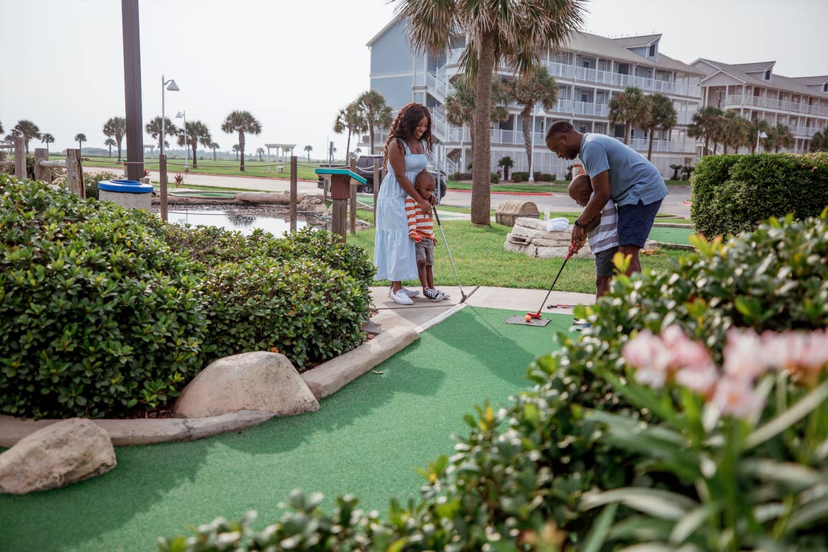A woman and young boy watch as a man helps a young boy tee up on a mini golf course outdoors.