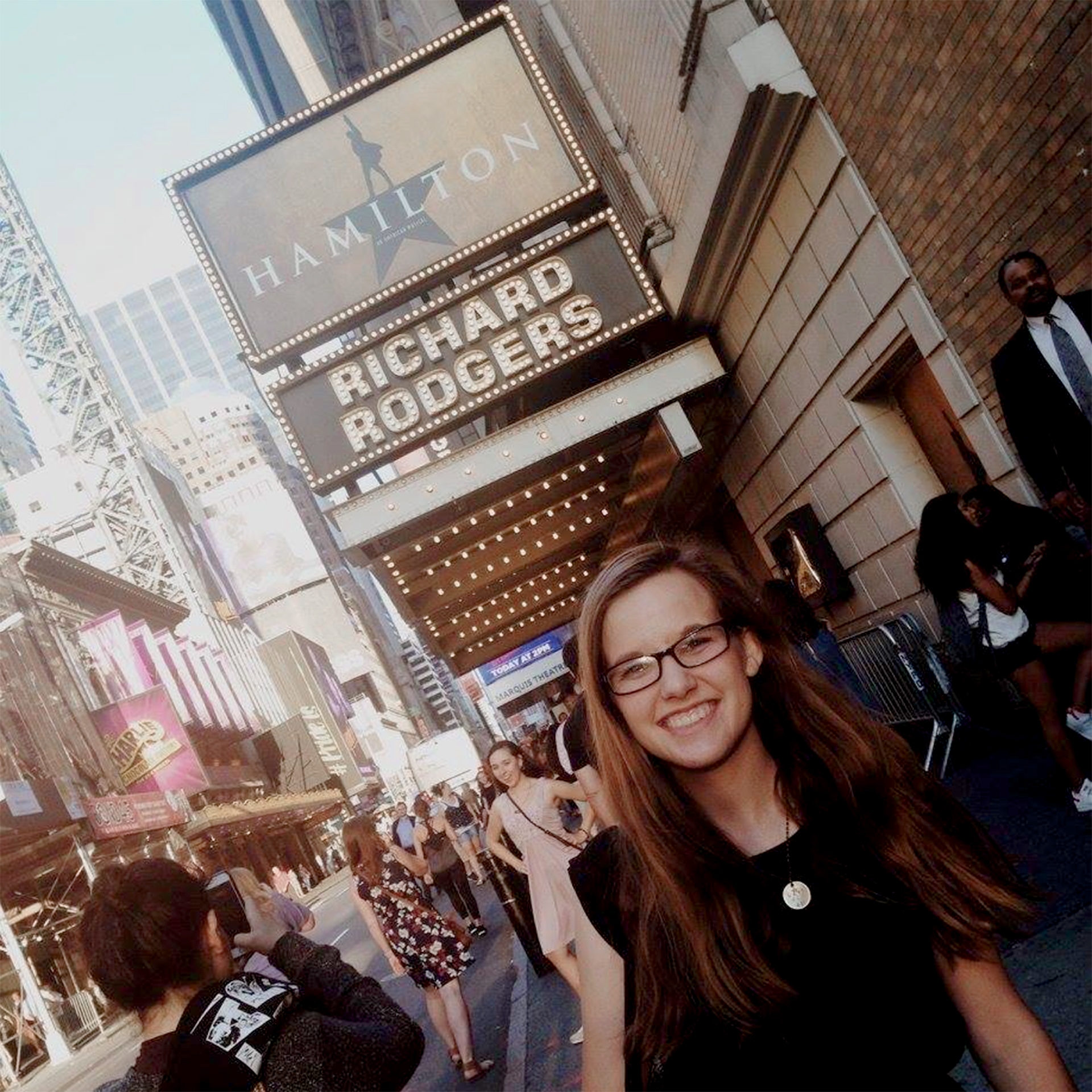 A caucasian woman with long ash-blonde hair wears a pair of black sunglasses and t-shirt under a marquee that reads, 'Hamilton, Richard Rodgers Theater' in New York City.