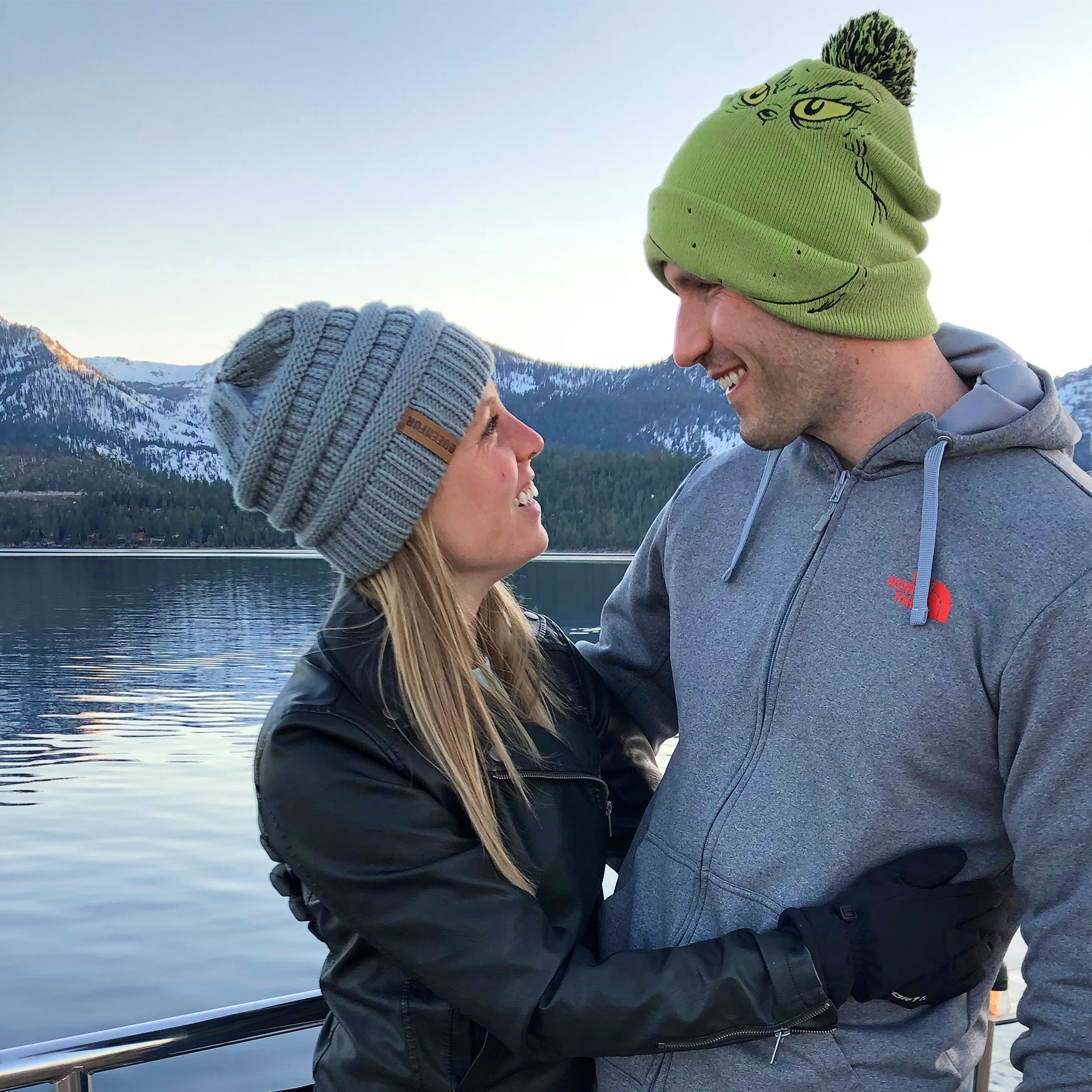 A woman (left) wears a grey beanie and black leather jacket and gloves while hugging a man (right) wearing a grey sip-up hoodie and green beanie.