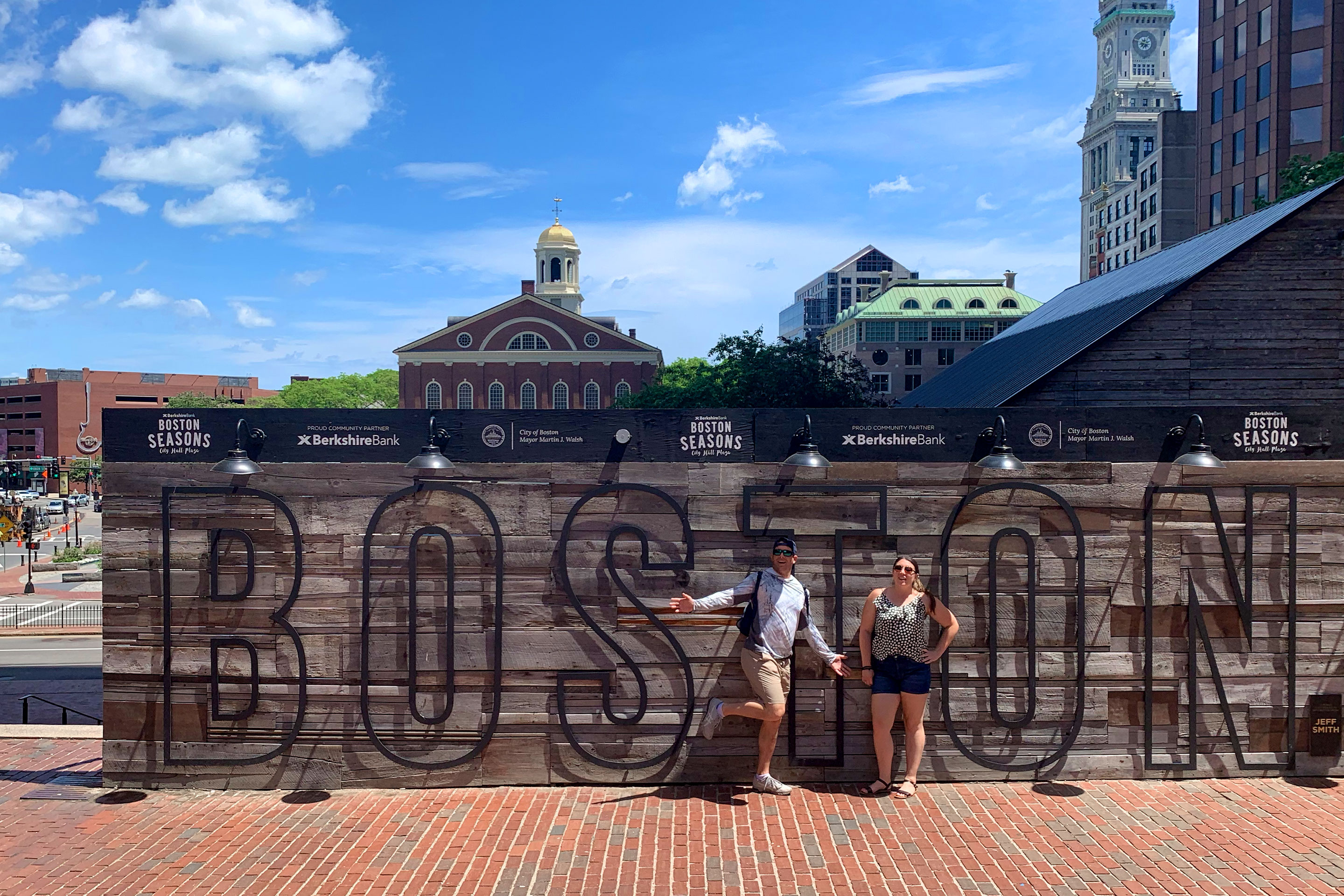 A man and woman stand near a shiplapped wall that reads 'BOSTON' in downtown Boston.