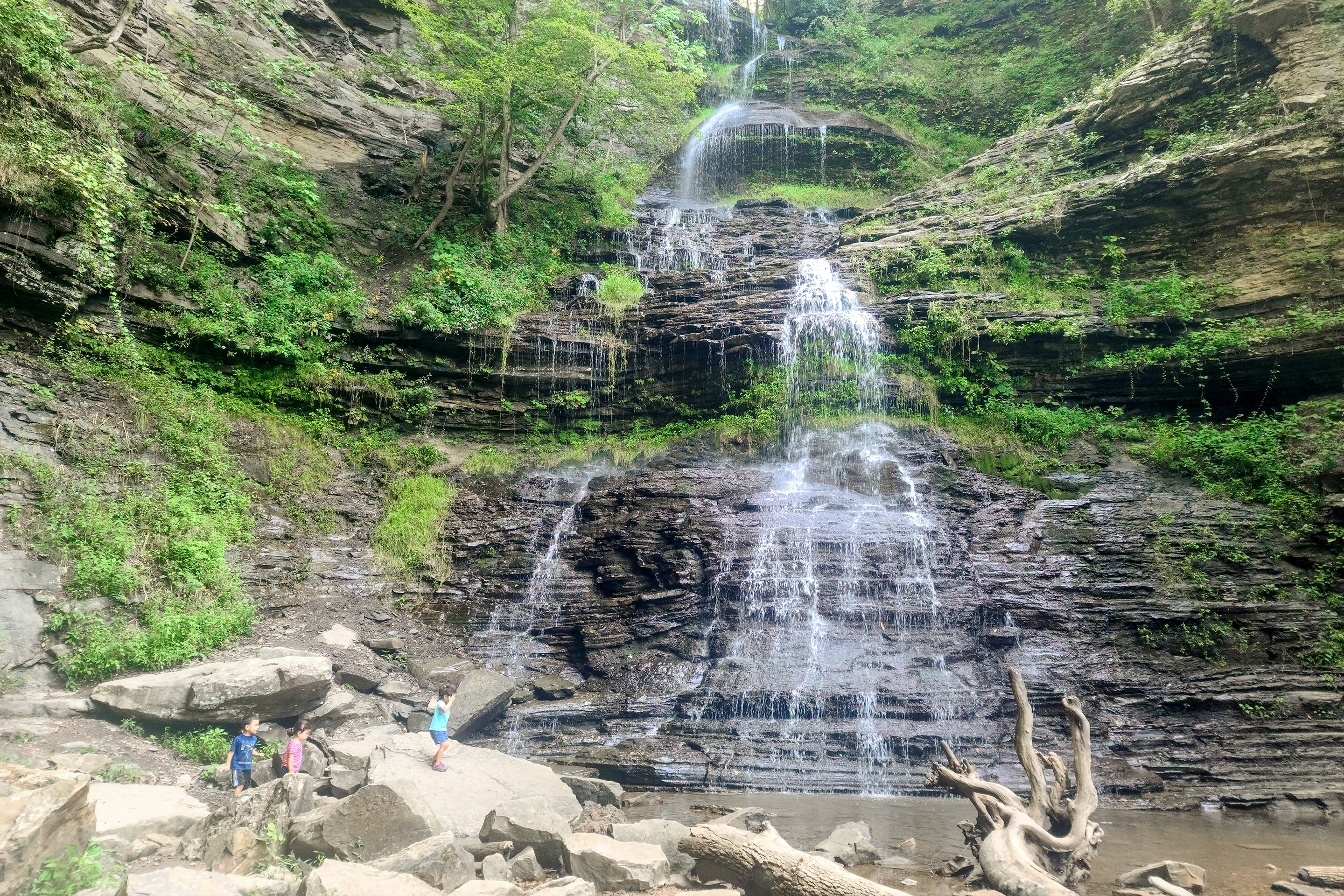 Three Asian Pacific Islander toddlers (left to right: Two girls and a boy) wear shorts and t-shirts while climbing rocks near a waterfall.