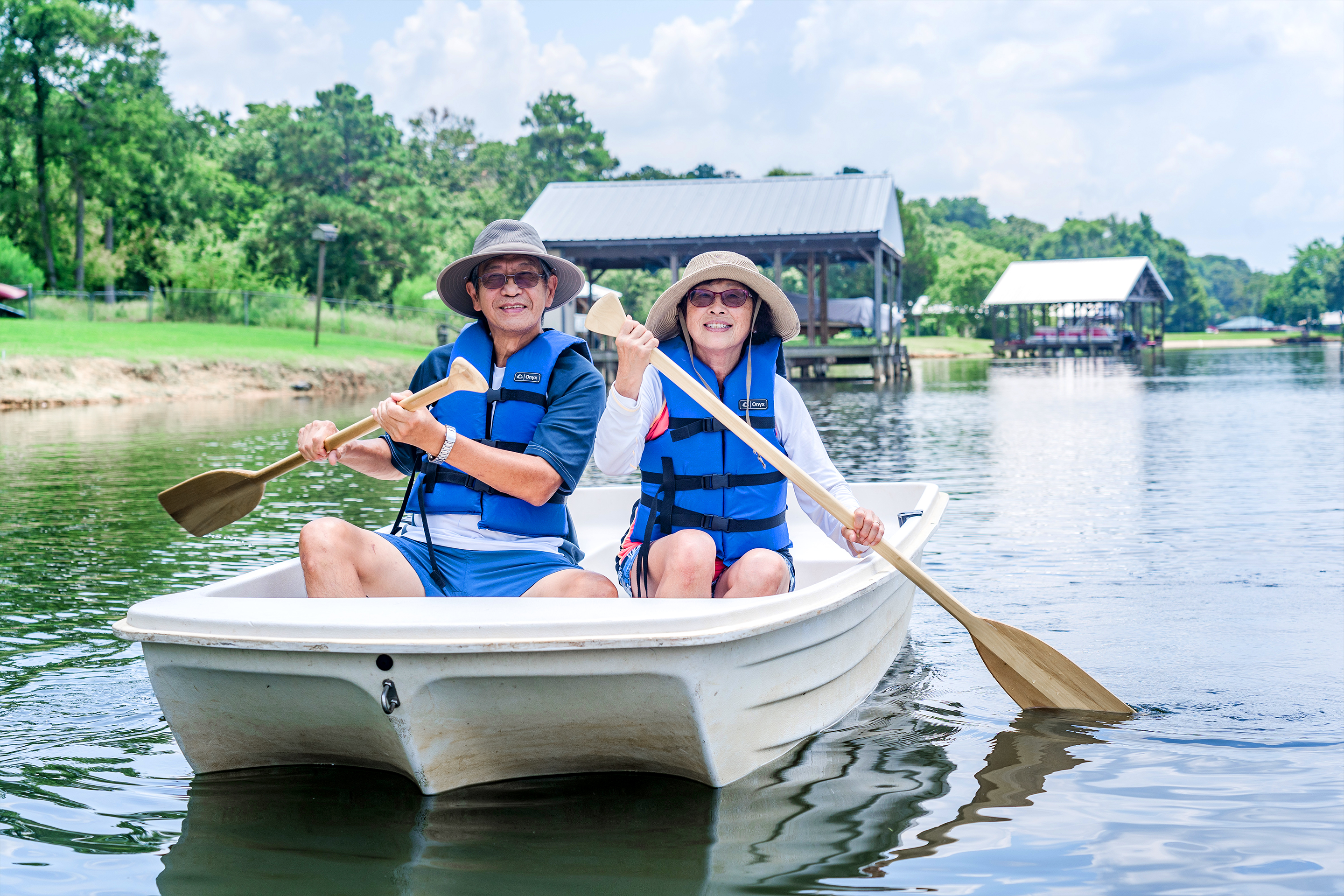An older Asian male (left) and female (right) wear blue life vests, sunglasses and various hats while riding on a white rowboat in a lake.