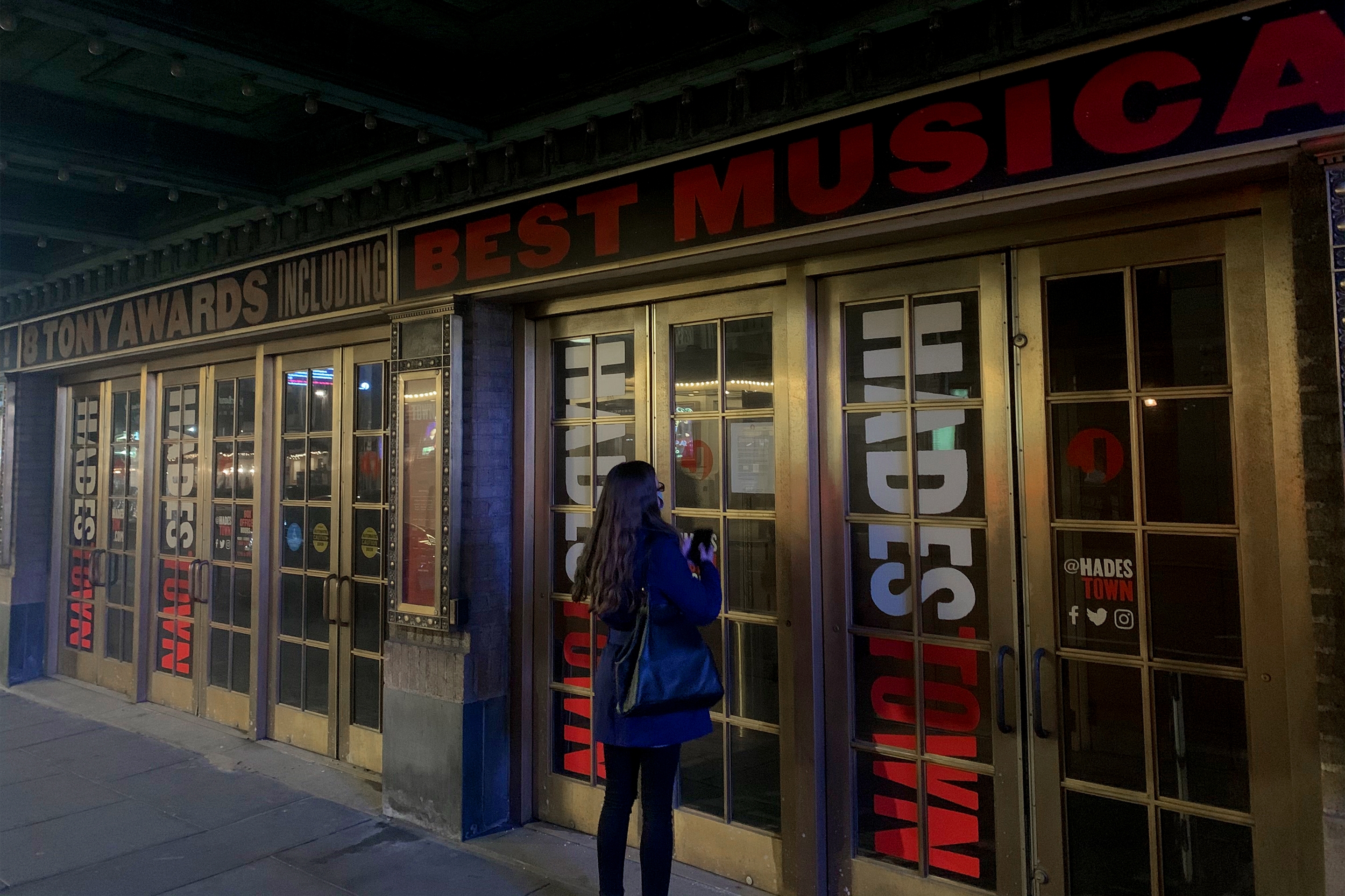 A caucasian woman with long ash-blonde hair wears a safety mask and blue jacket standing outside of a theater vestibule in New York City.