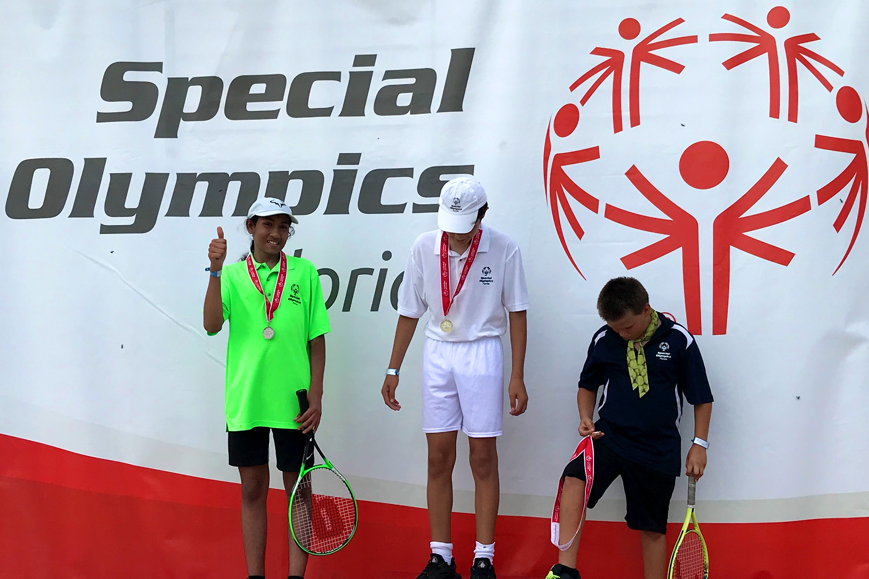 Special Olympic Athlete, Roan Luallen, wears a neon green t-shirt and black shorts while holding a tennis racket and wearing a medal along with two other competitors at the Special Olympics in 2019.