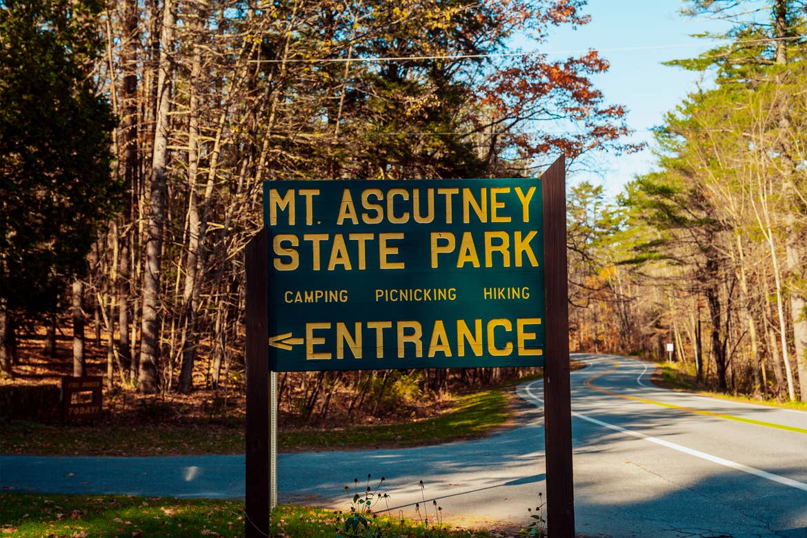 A green, wooden carved state park sign with yellow lettering placed on the side of a road surrounded by fall foliage.