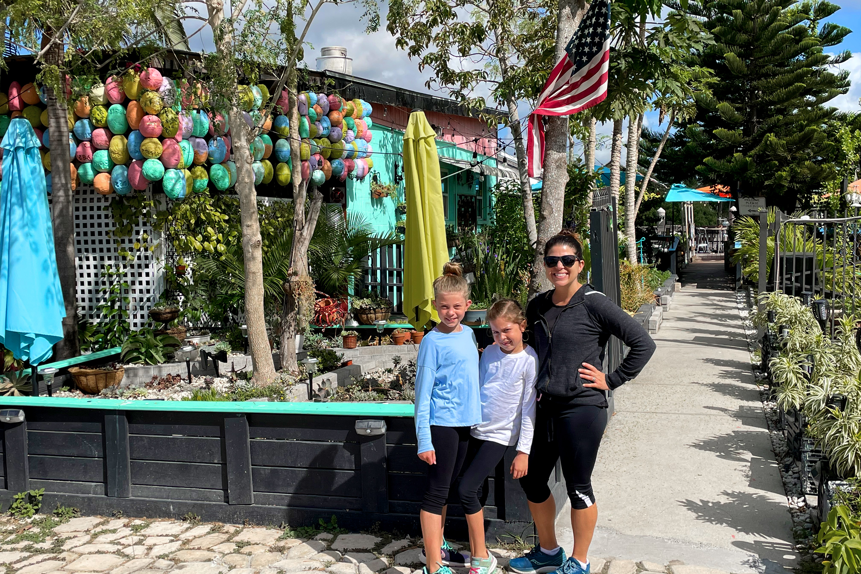 A caucasian woman (right) wearing a black shirt and sunglasses poses next to two young caucasian girls, one wearing a blue t-shirt (left) and another (middle) wearing a long sleeve t-shirt near a succulent garden.