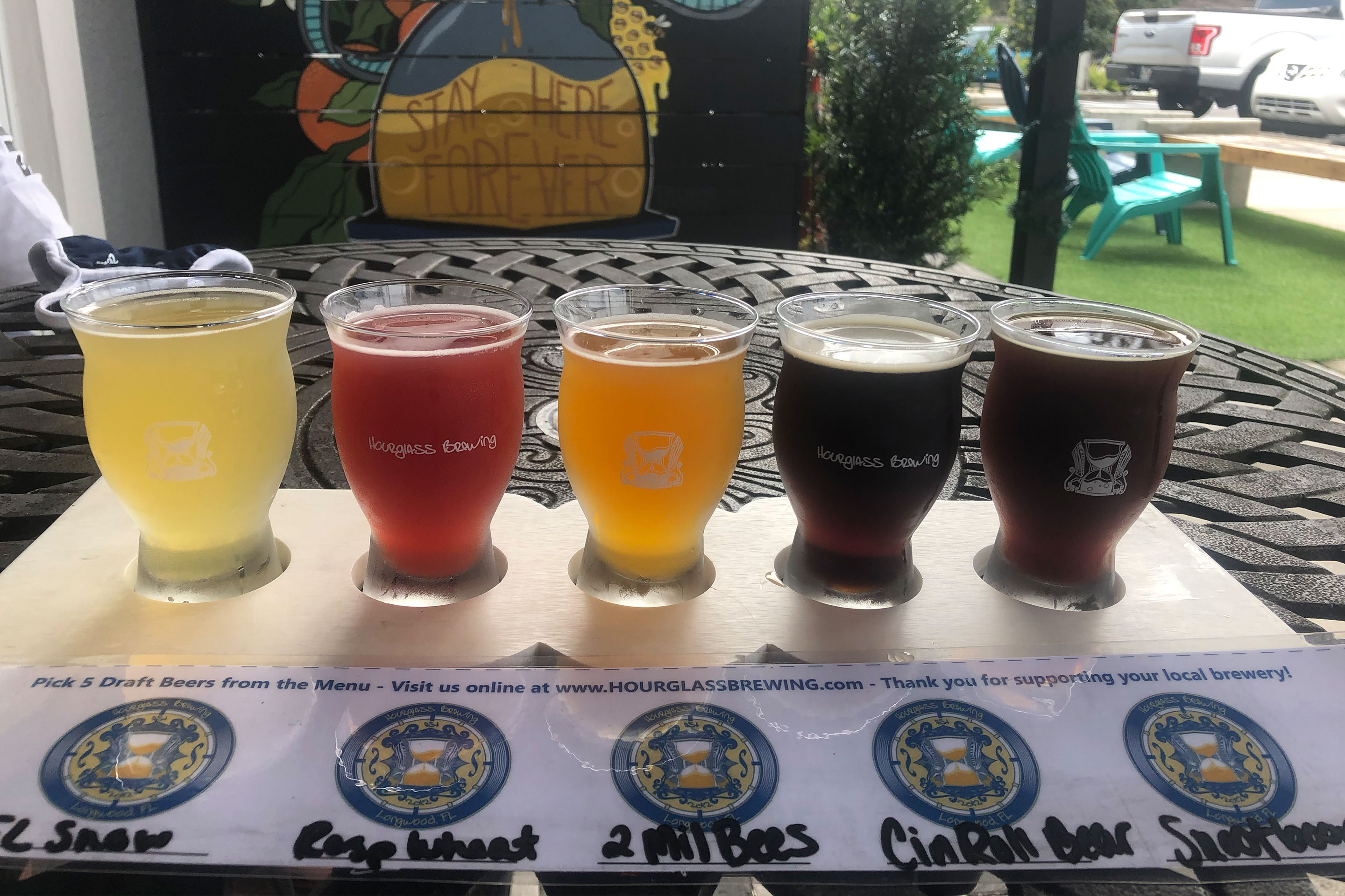 A beer flight with a label that reads abbreviations, '1. FL Snow 2. Raspberry Wheat 3. 2 Million Bees 4. Cinnamon Roll Bear 5. Snootboops from 'Hourglass Brewing.'