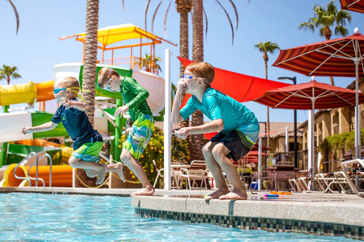 Three young boys wearing all their swimwear, jump into the pool near our water slides at our Scottsdale Resort.