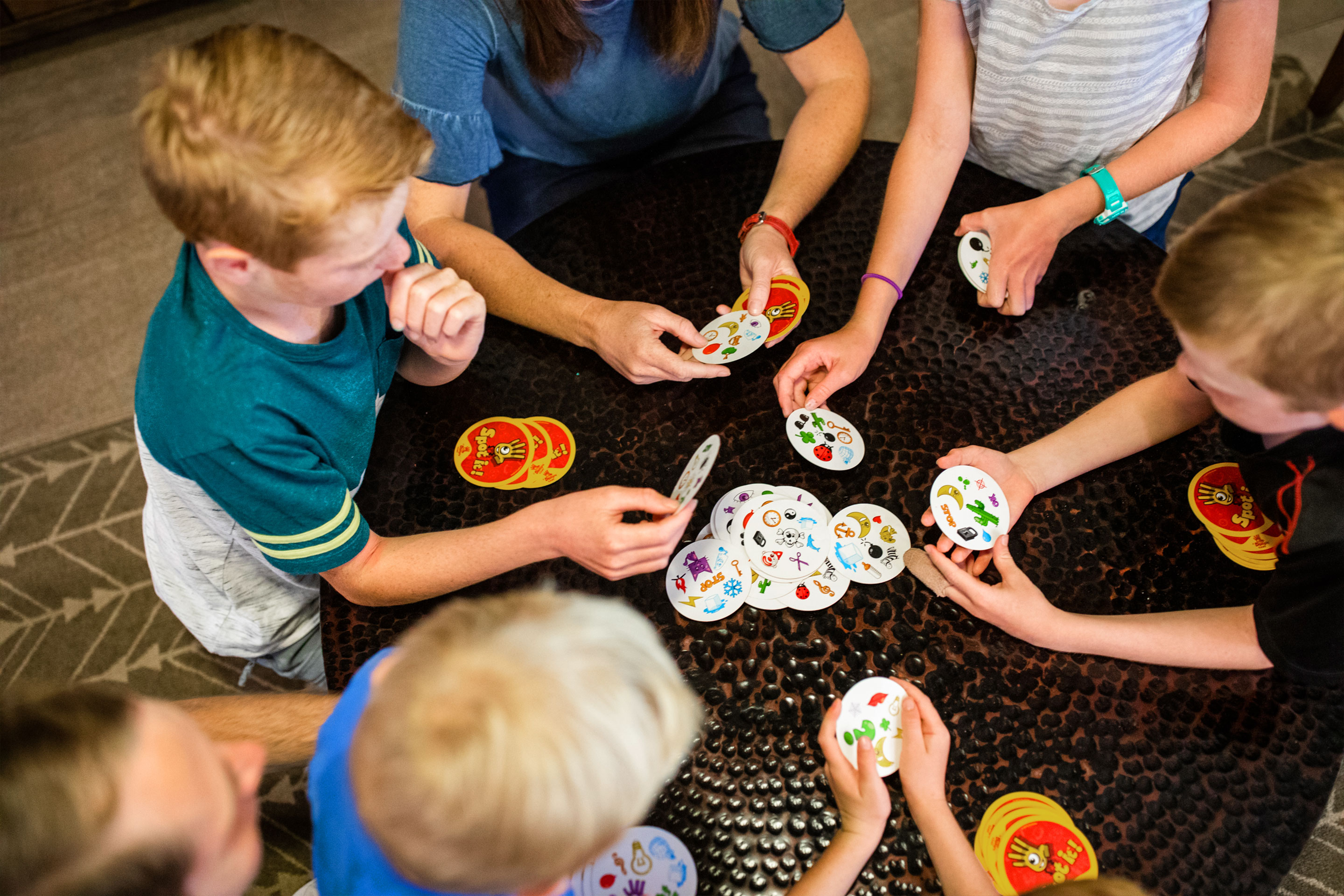 Several children and two adults play a card game on a coffee table.