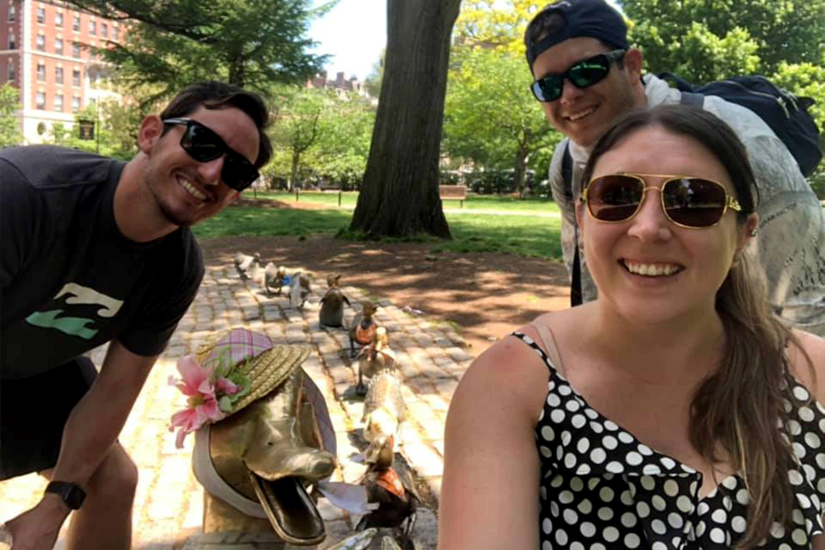 Two men and a woman pose next to several bronze statues of a duck and her ducklings wearing bonnets.