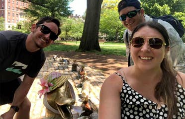 Two men and a woman pose next to several bronze statues of a duck and her ducklings wearing bonnets.
