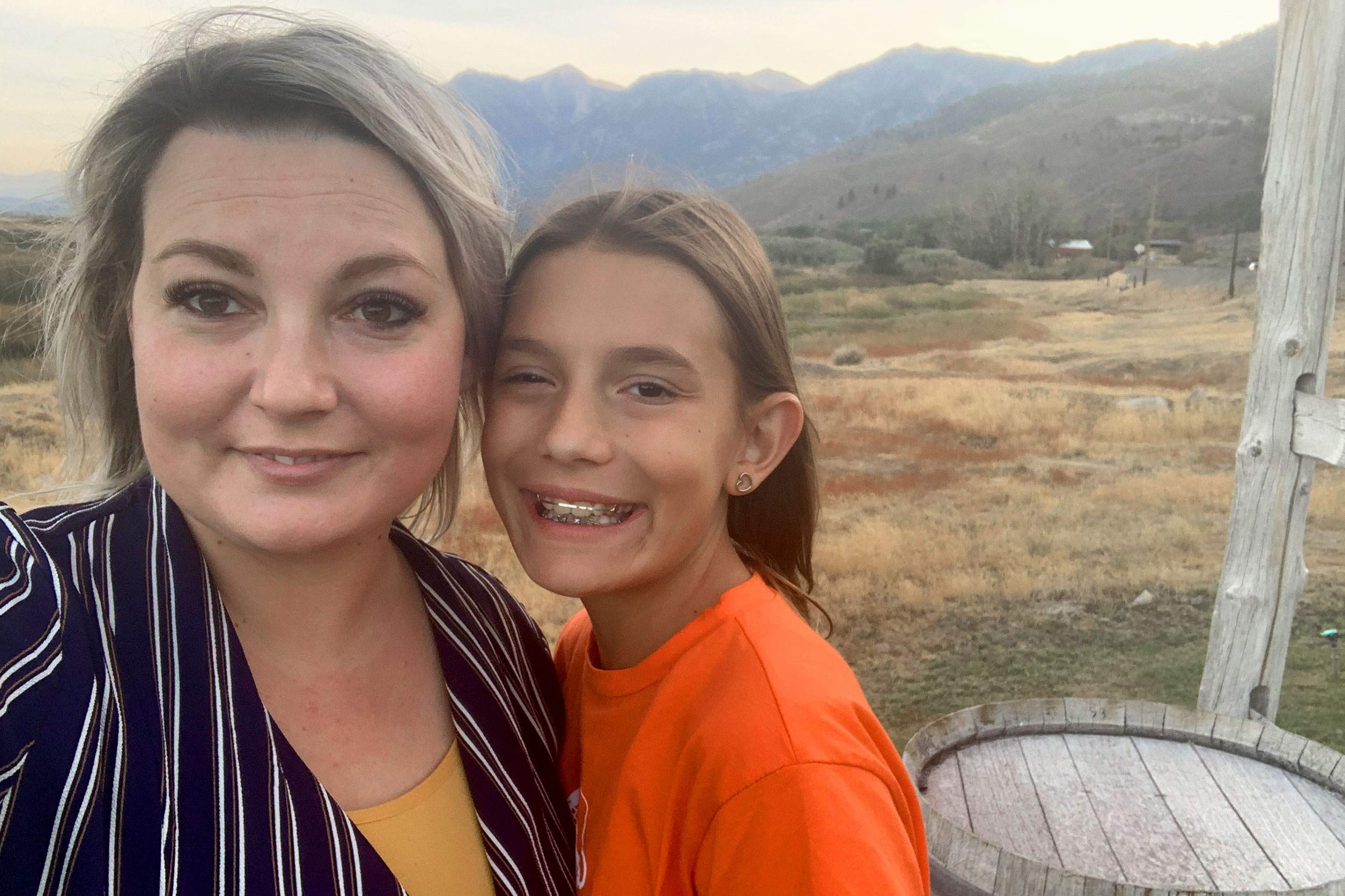 A woman in a striped, black blouse stands next to a young girl in an orange t-shirt in front of a mountain range.