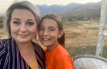 A woman in a striped, black blouse stands next to a young girl in an orange t-shirt in front of a mountain range.