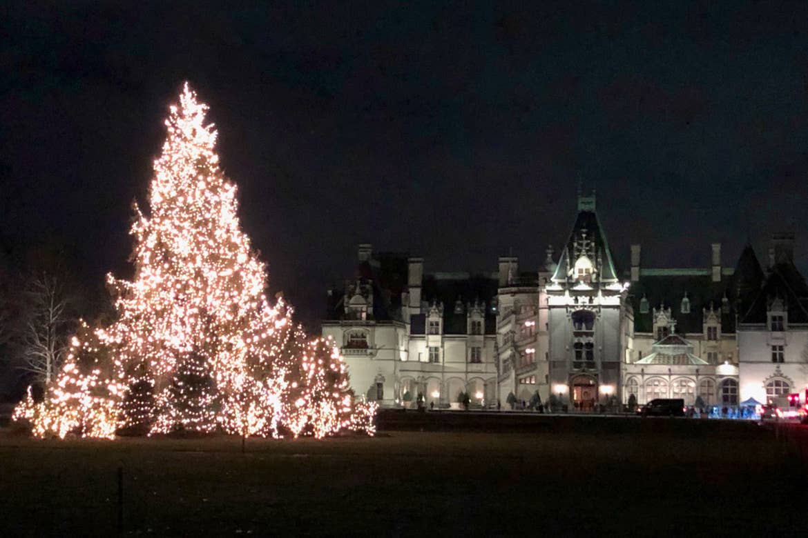 The exterior of the Biltmore Estate at night with a large, string light covered Christmas tree on the front lawn.
