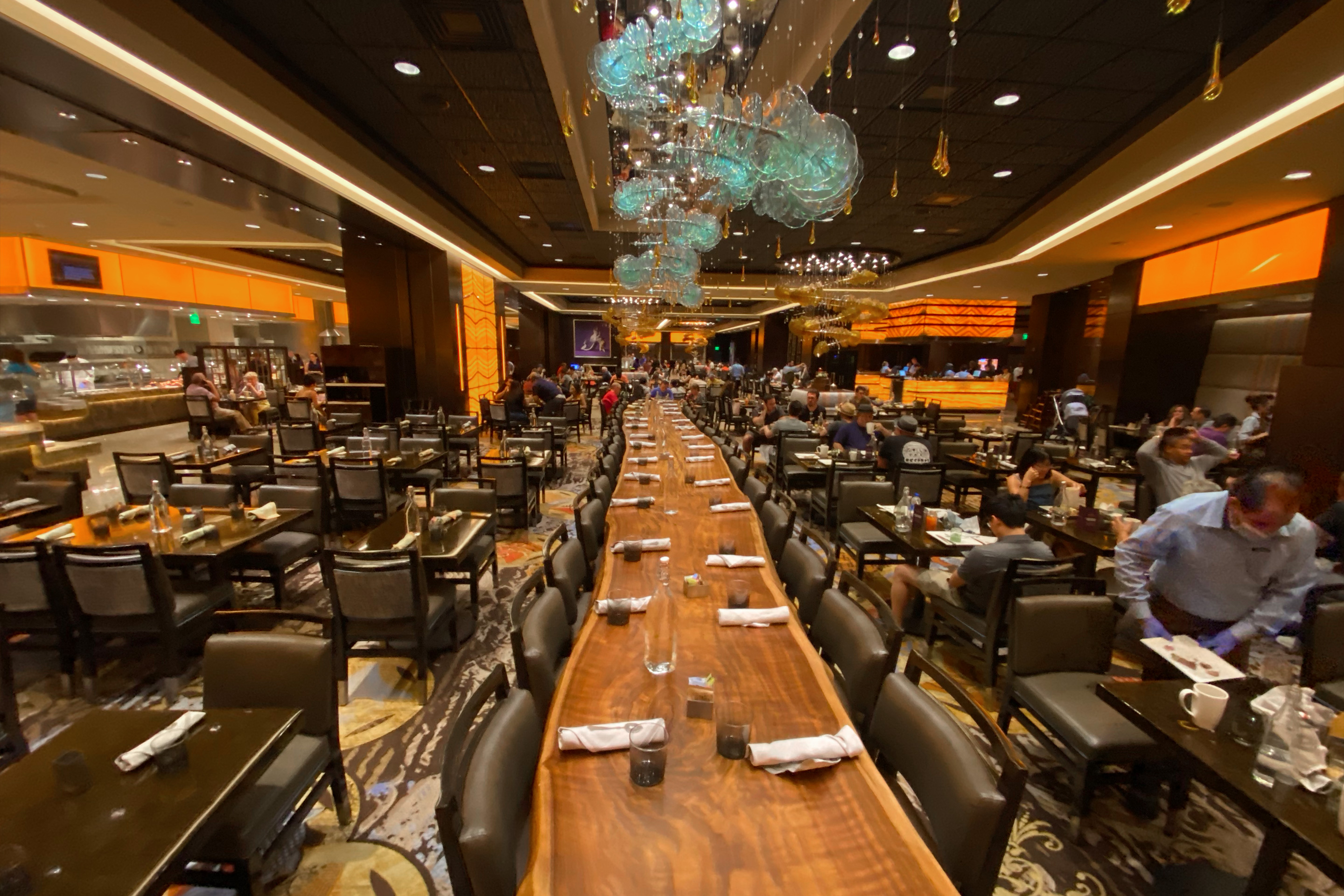 A long wooden communal table with place settings for and multiple chairs surrounded by other tables for diners.