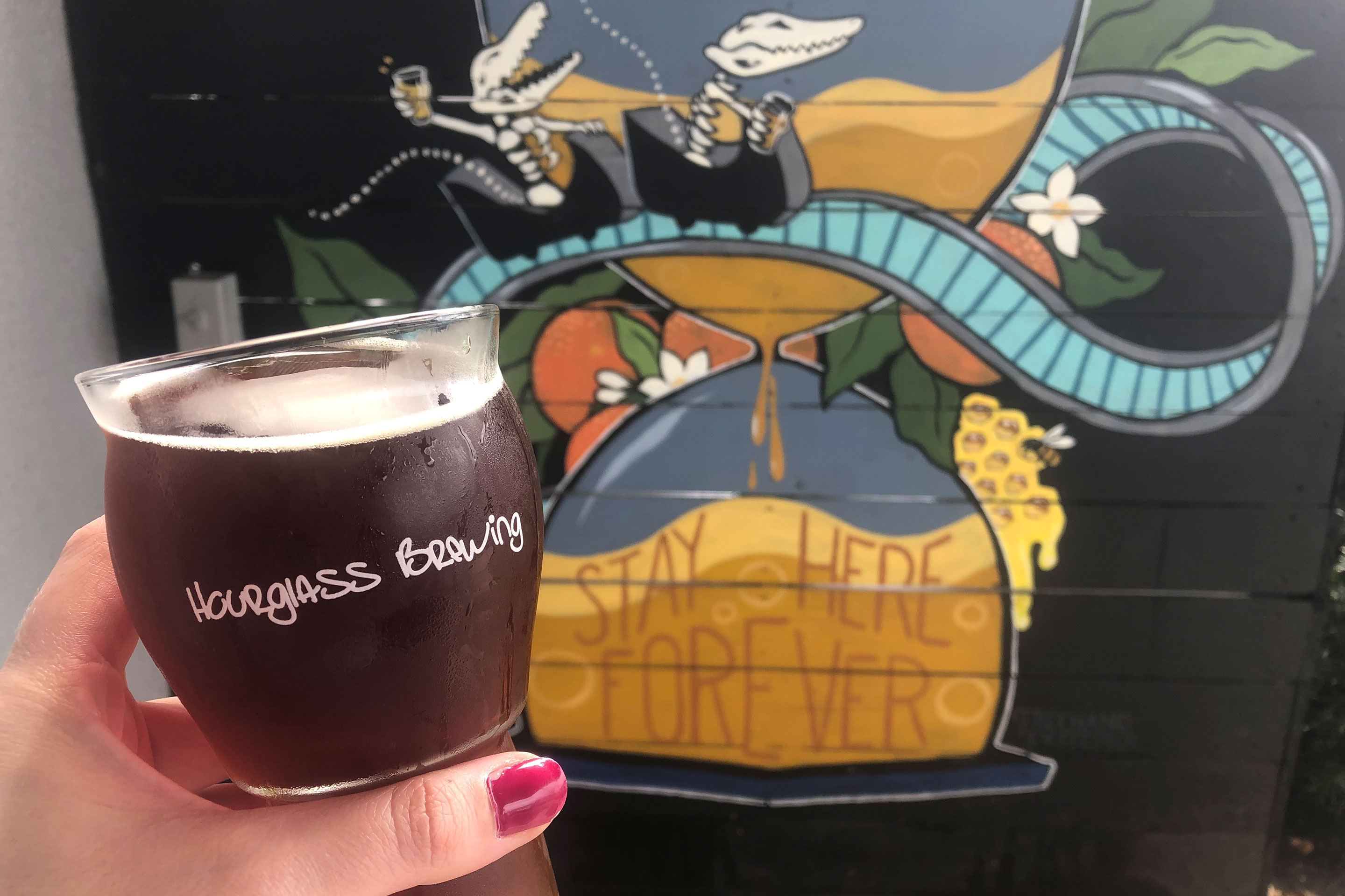 A hand holds a beer glass with the 'Hourglass Brewing' logo on it in front of a wall mural that reads, 'Stay Here Forever' at  'Hourglass Brewing.'