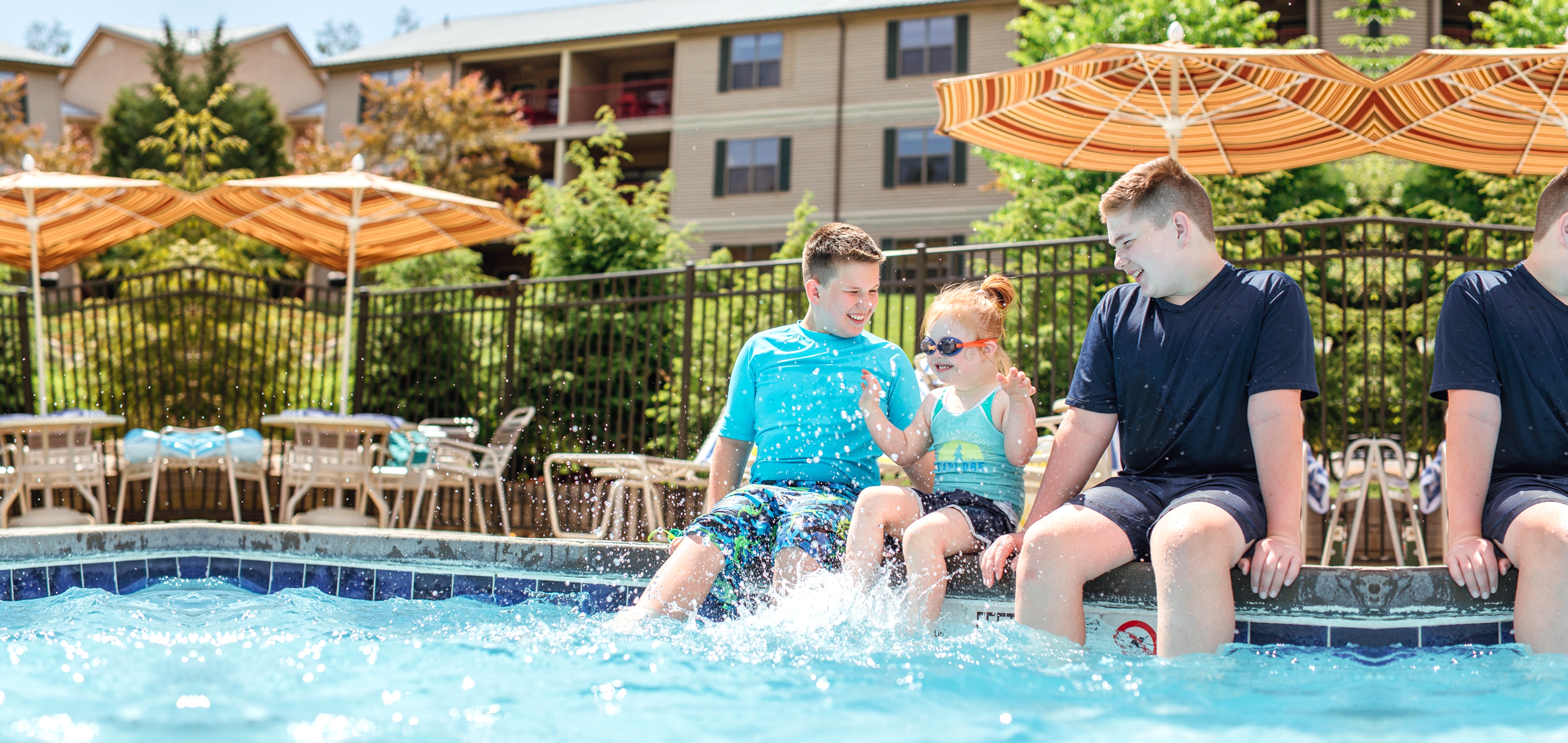 Two young caucasian tween boys (left and right) wearing swim shirts and shorts sit with their feet in a pool with a young caucasian girl (middle) wearing a swimsuit, swimming shorts and goggles as she splashes her feet.