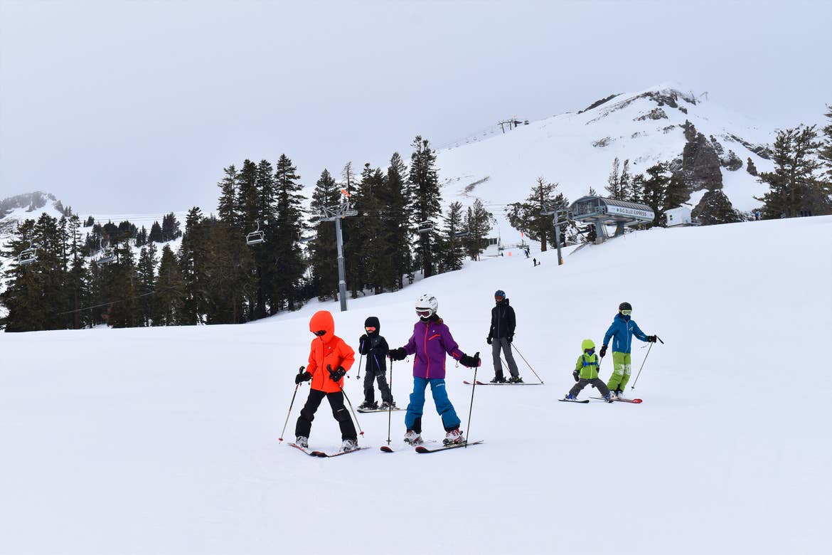 Five children and a man wearing ski apparel make their way down a snowy mountain.