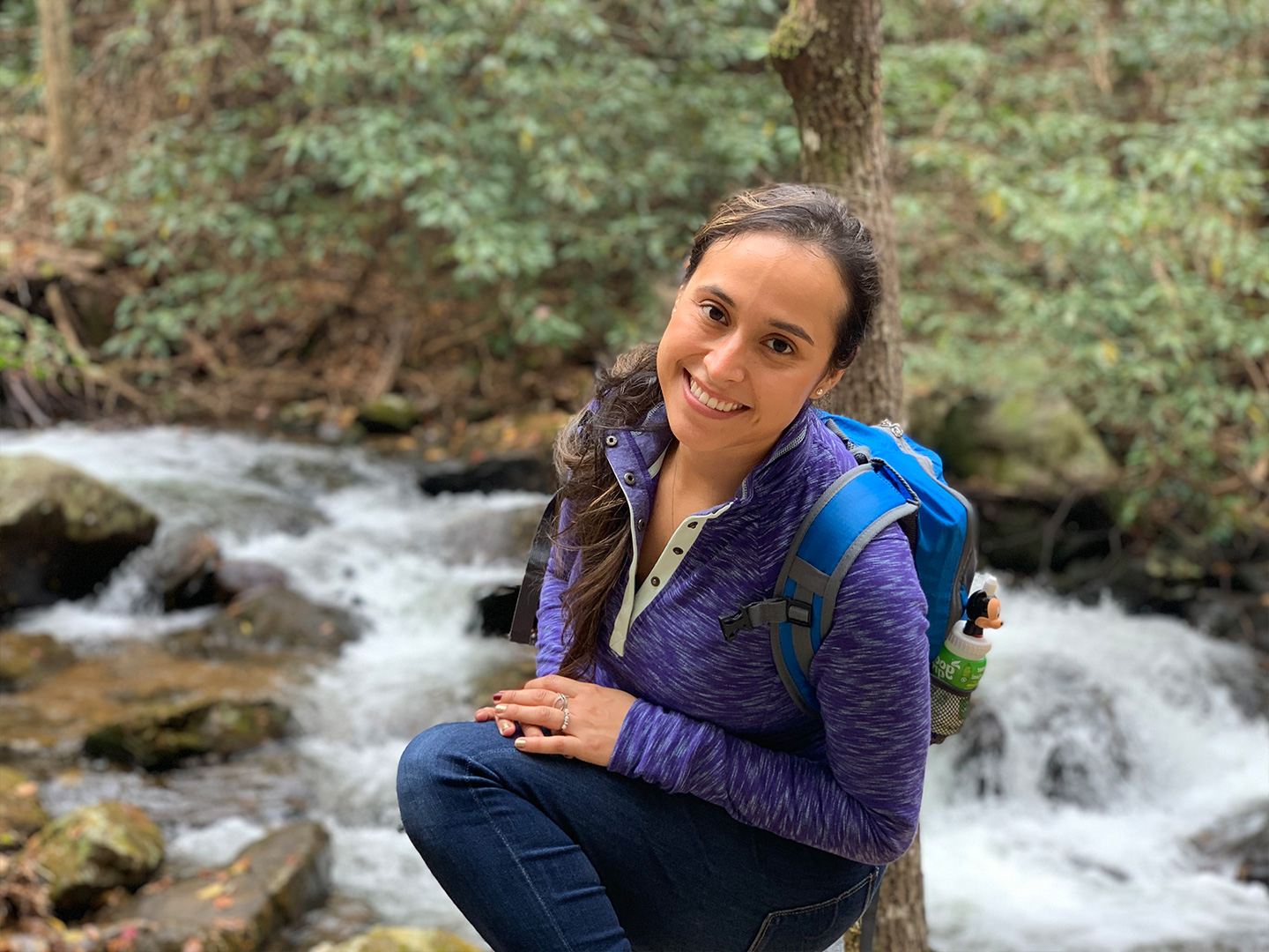 Featured author, Andrea Beltran, poses near a creek wearing a blue hiking backpack and purple pull-up sweater.