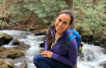 Featured author, Andrea Beltran, poses near a creek wearing a blue hiking backpack and purple pull-up sweater.