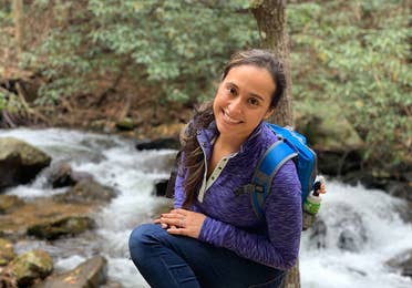 Featured author, Andrea Beltran, poses near a creek wearing a blue hiking backpack and purple pull-up sweater.