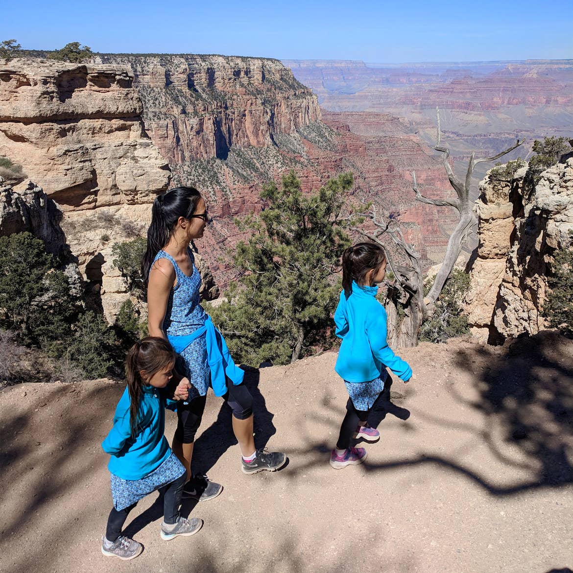 Two young girls and a woman dressed in blue walk on a trail alongside the Grand Canyon.