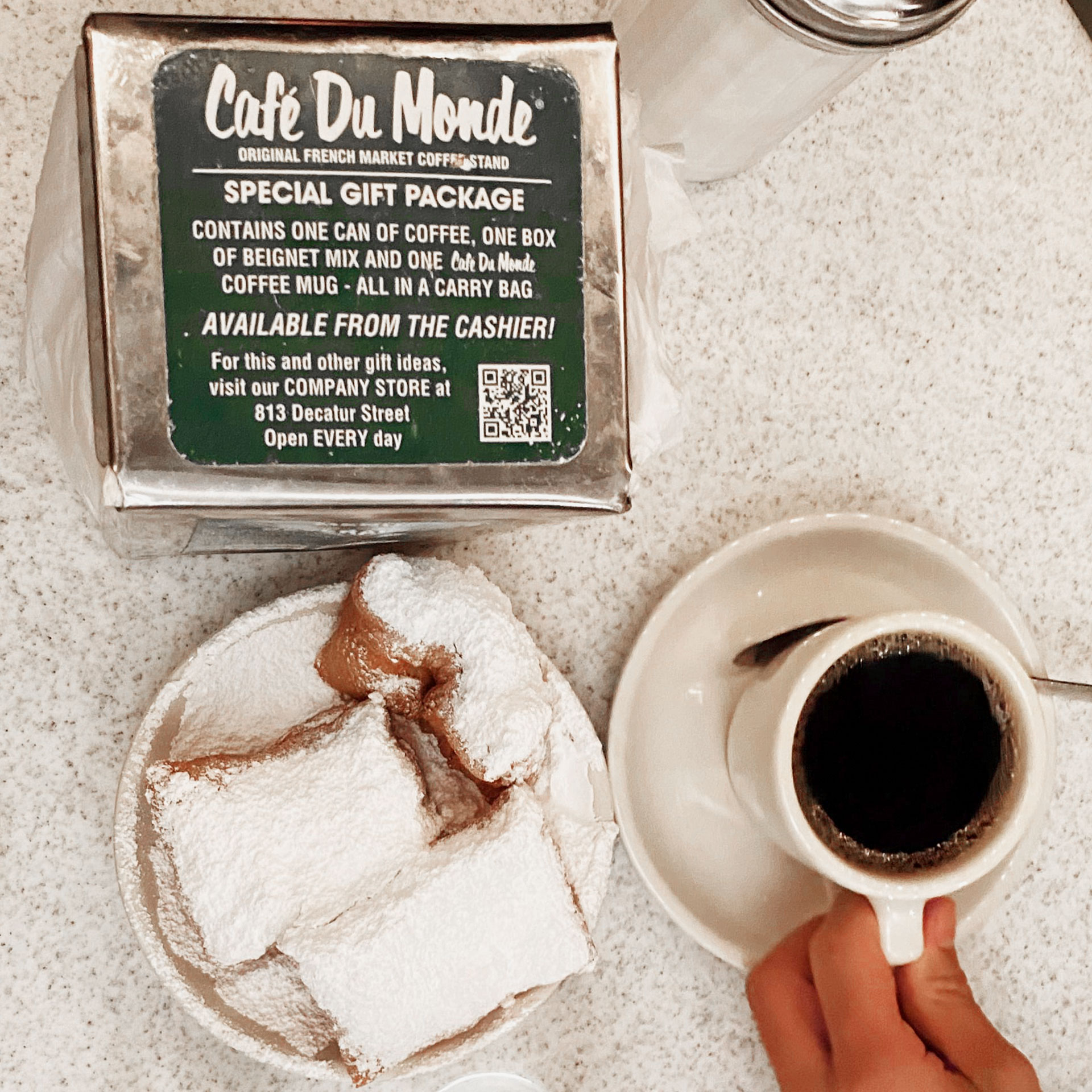 A Cafe Du Monde napkin box with beignets placed on a table with some coffee on white plates.