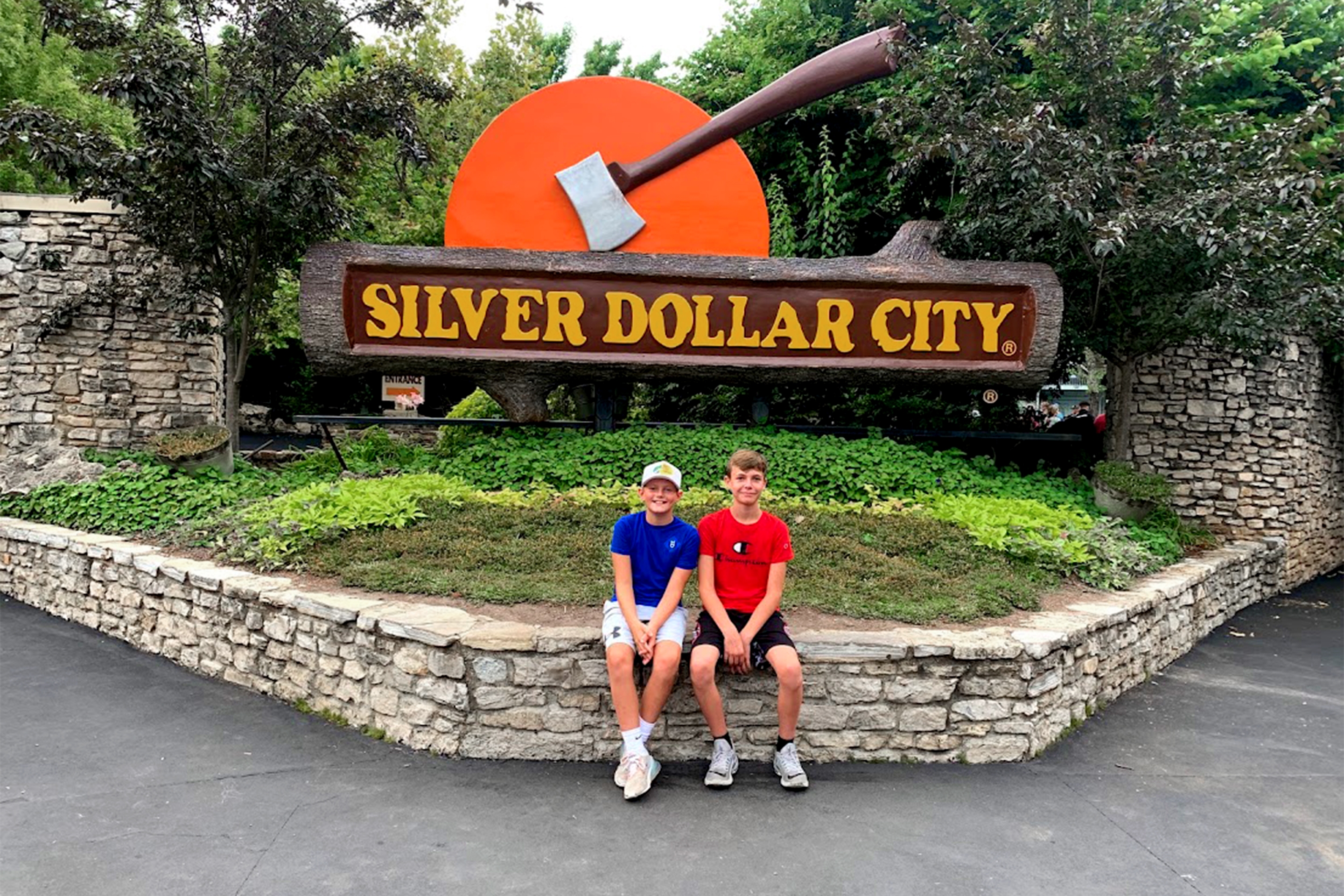 Two boys wearing t-shirts, basketball shorts and sneakers sit on a stone wall in front of some landscaping and a giant log sign that reads, 'Silver Dollar City.'