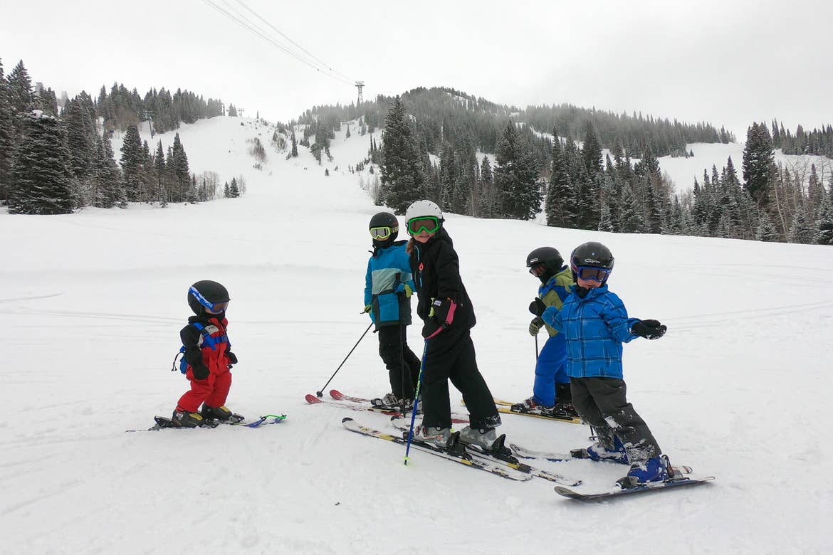 Five young children wear winter apparel, skiing helmets, skiing goggles and a set of skis while standing on a snowy slope.
