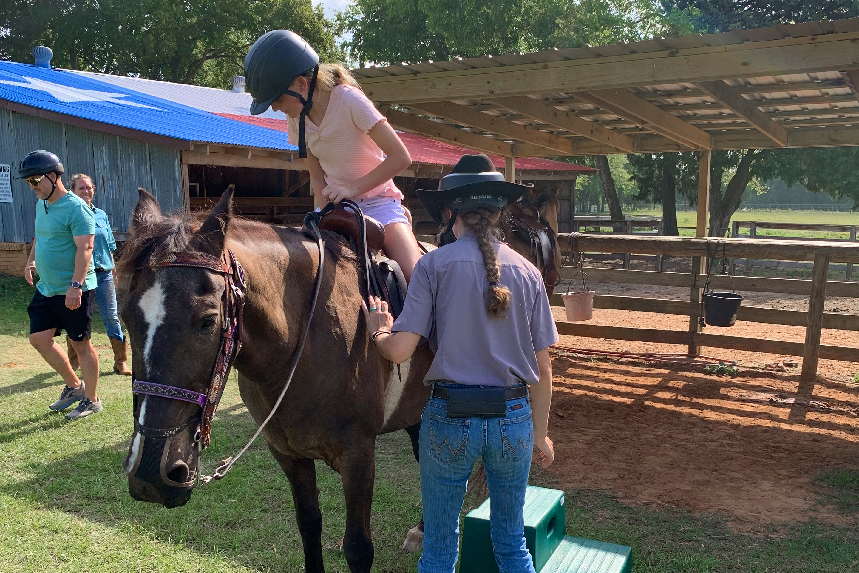 A caucasian girl (left) mounts a horse with the assistance of a handler.