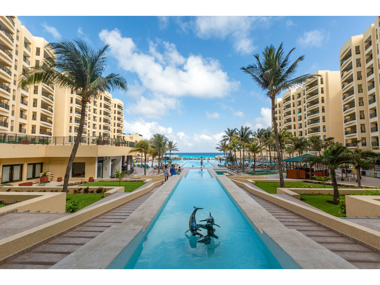 Resort view with a long pool featuring dolphin sculptures, lined with palm trees, leading toward the beach.