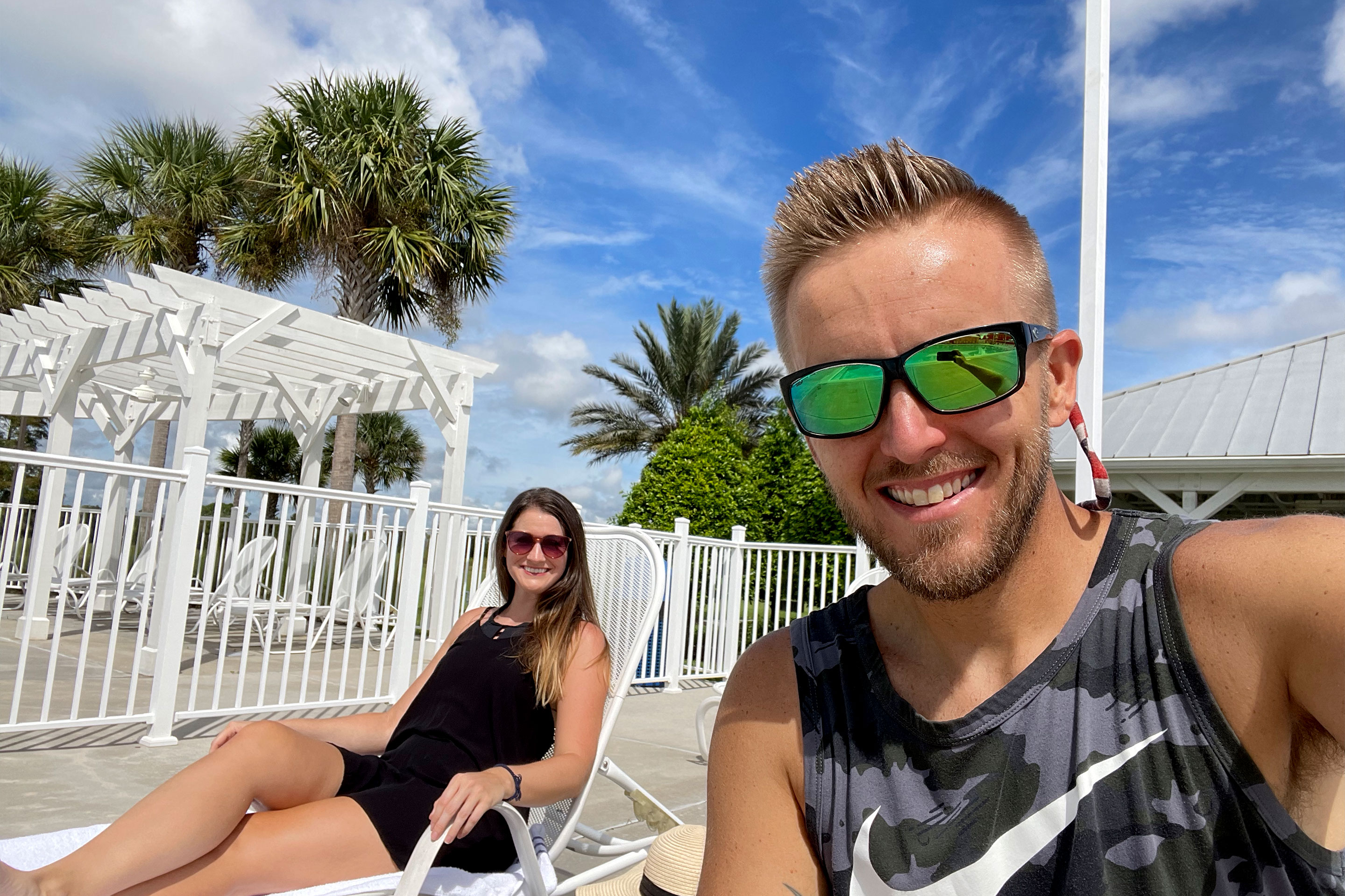 A woman in black (left) sits on a lounge chair next to a man in a camo tank (front) poolside under a blue sky.