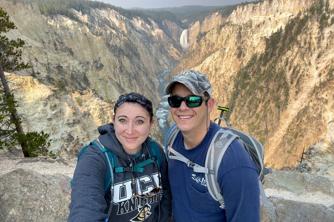 A woman in a dark grey hoodie with backpack and a man wearing a dark, long sleeve shirt, sunglasses, baseball cap and backpack stand in front of the Yellowstone Waterfall.