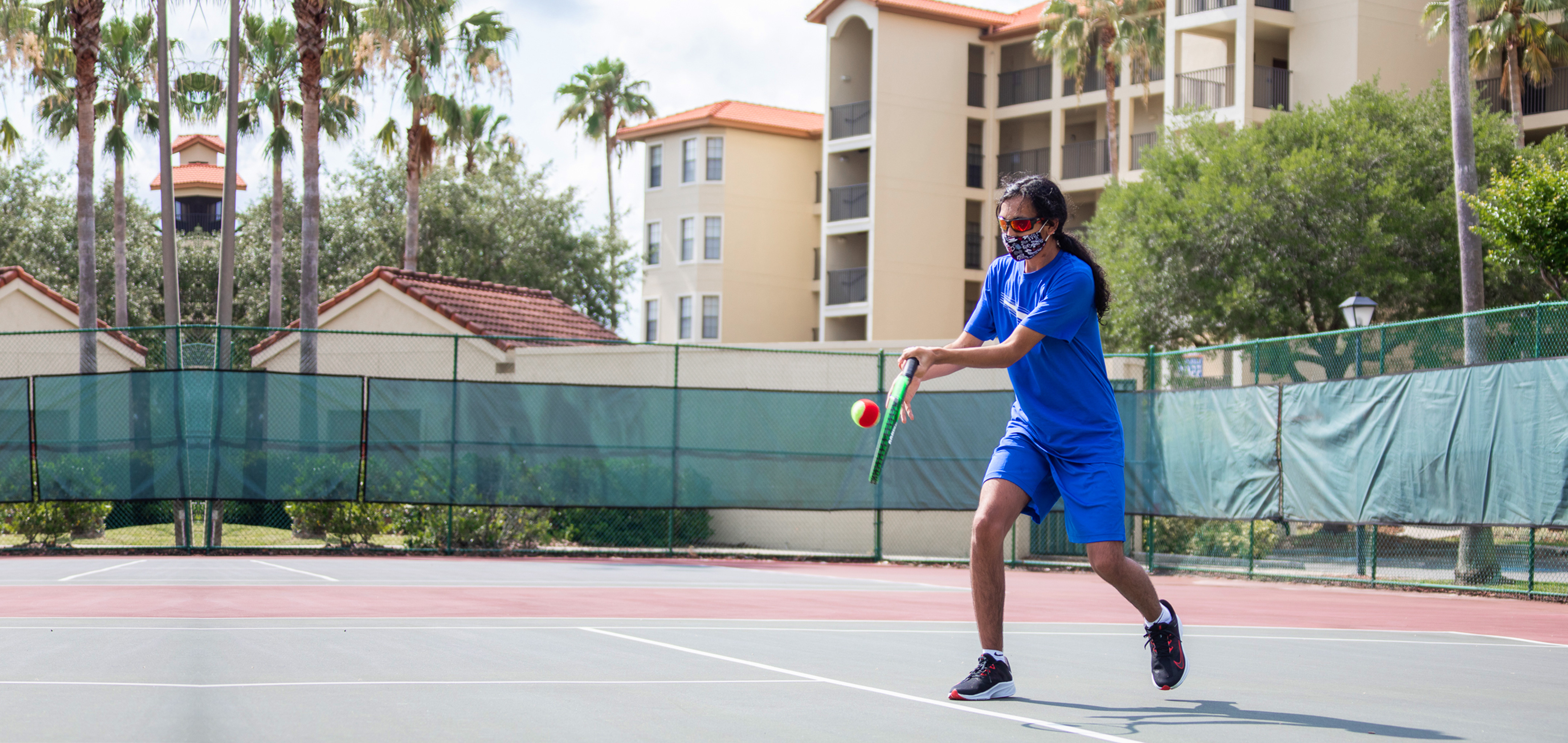 Special Olympic Athlete, Roan Luallen, plays tennis wearing a blue t-shirt and shorts with a safety mask and sunglasses on the courts of our Orange Lake Resort located in Orlando, Florida.