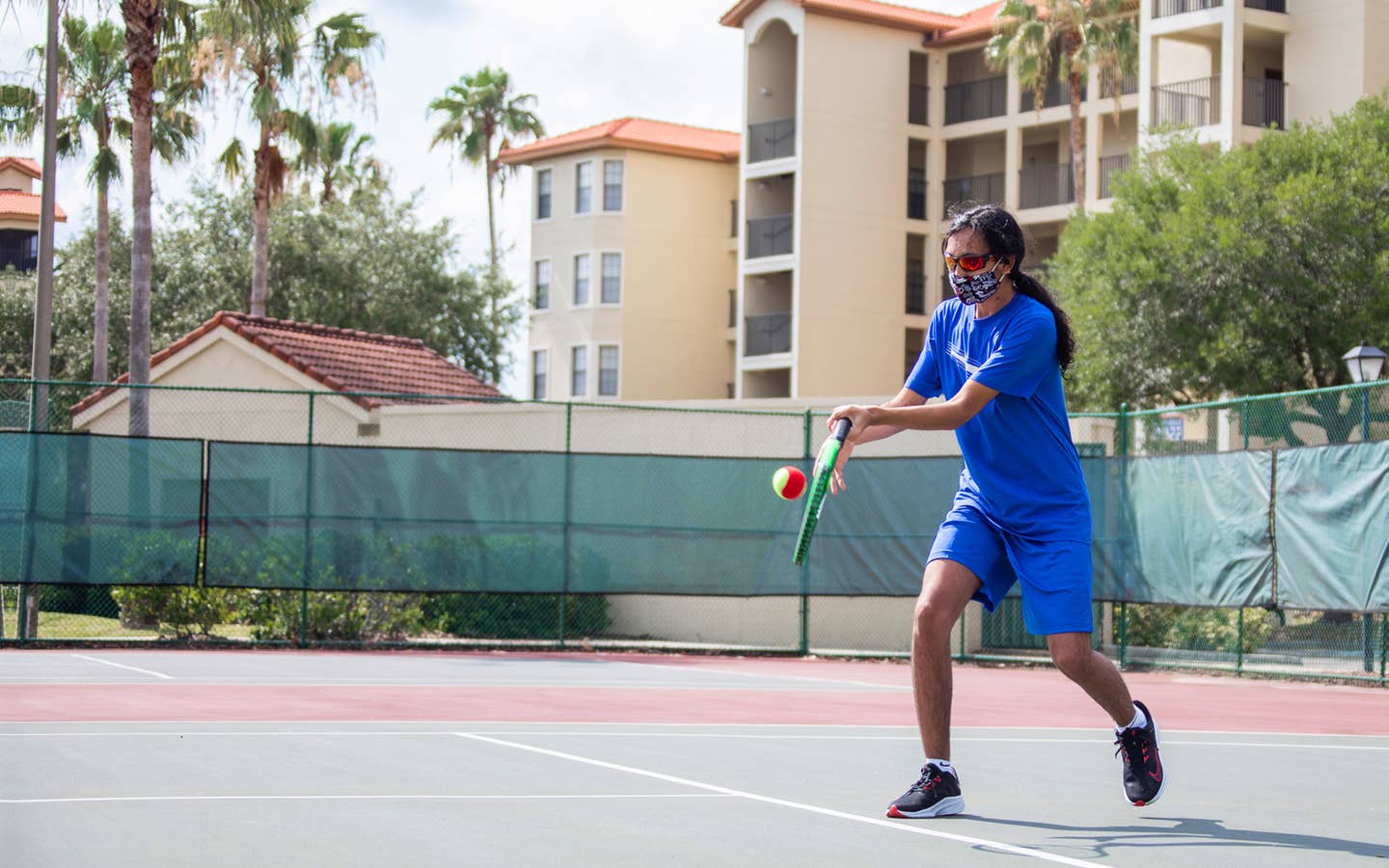 Special Olympic Athlete, Roan Luallen, plays tennis wearing a blue t-shirt and shorts with a safety mask and sunglasses on the courts of our Orange Lake Resort located in Orlando, Florida.