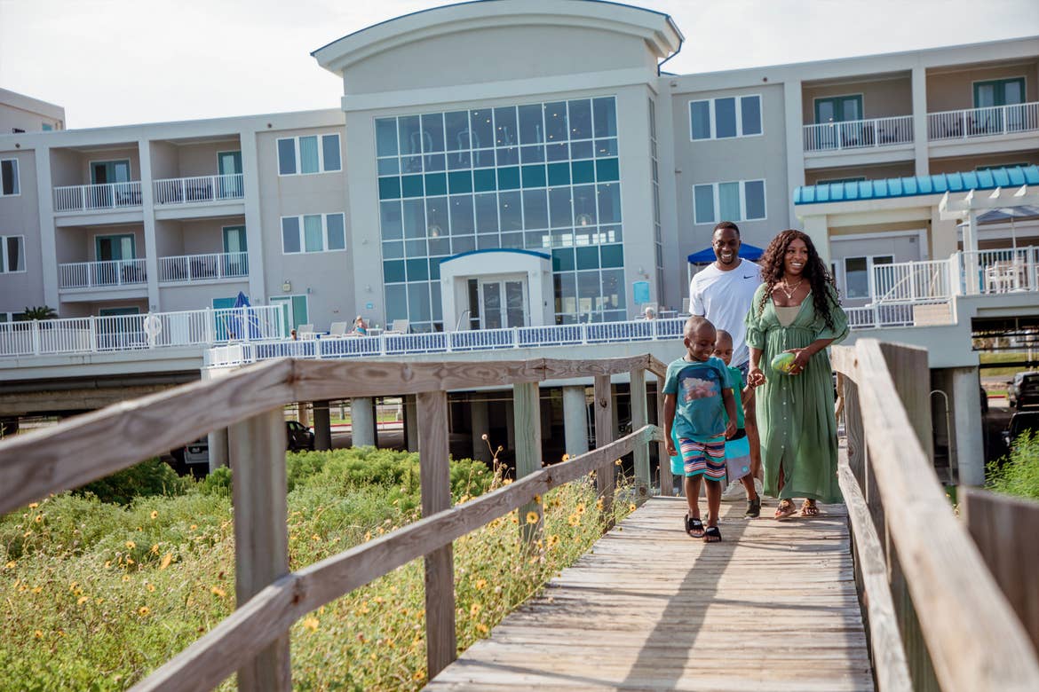 A man, woman and two young boys walk a wooden boardwalk from a resort exterior.