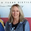 A blonde, caucasian woman stands in front of a Texas distillery vinyl sign while wearing a grey zip-up hoodie with a black t-shirt.