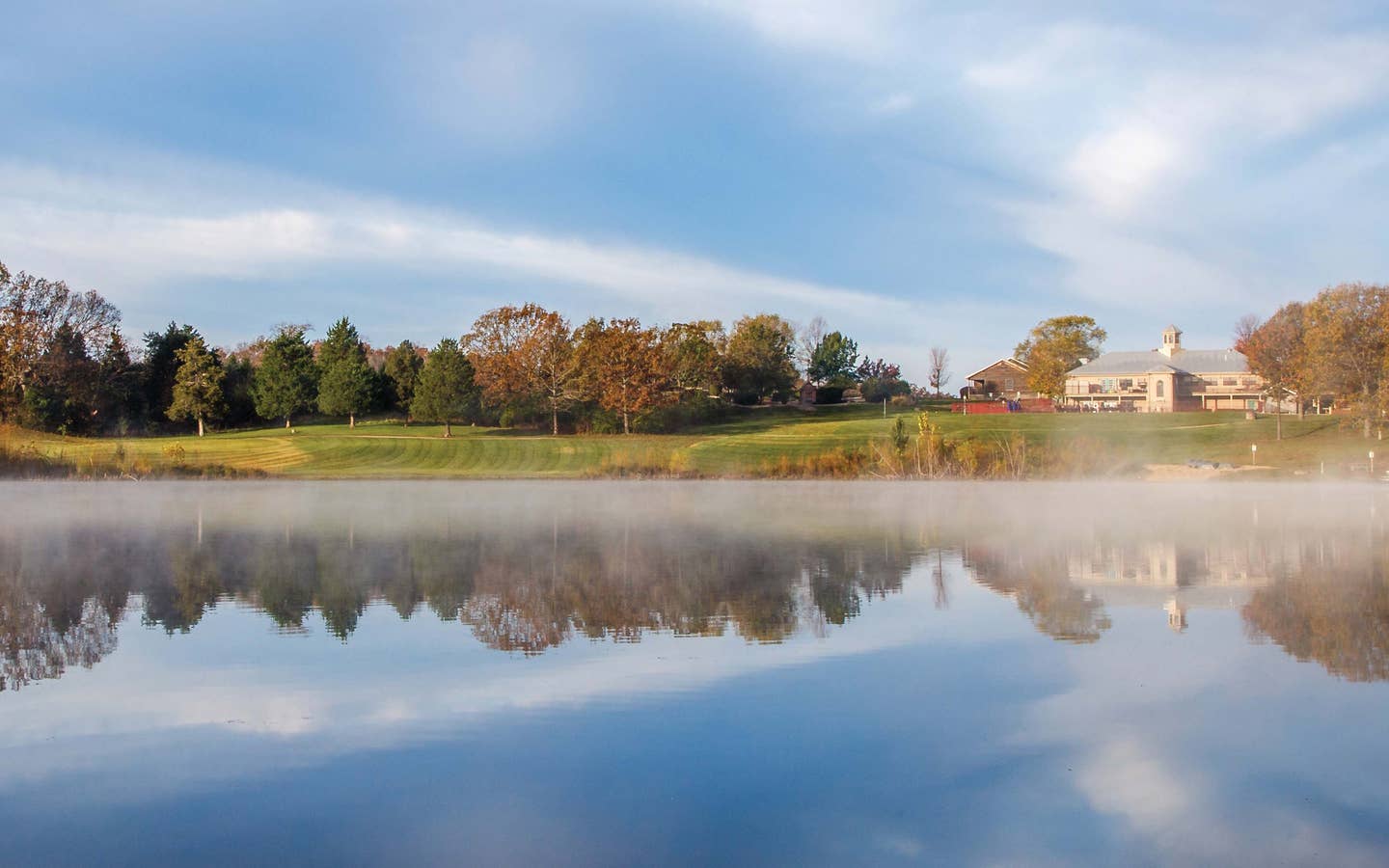 Lake with fog rolling on top and view of Timber Creek Resort property building and trees in De Soto, Missouri.