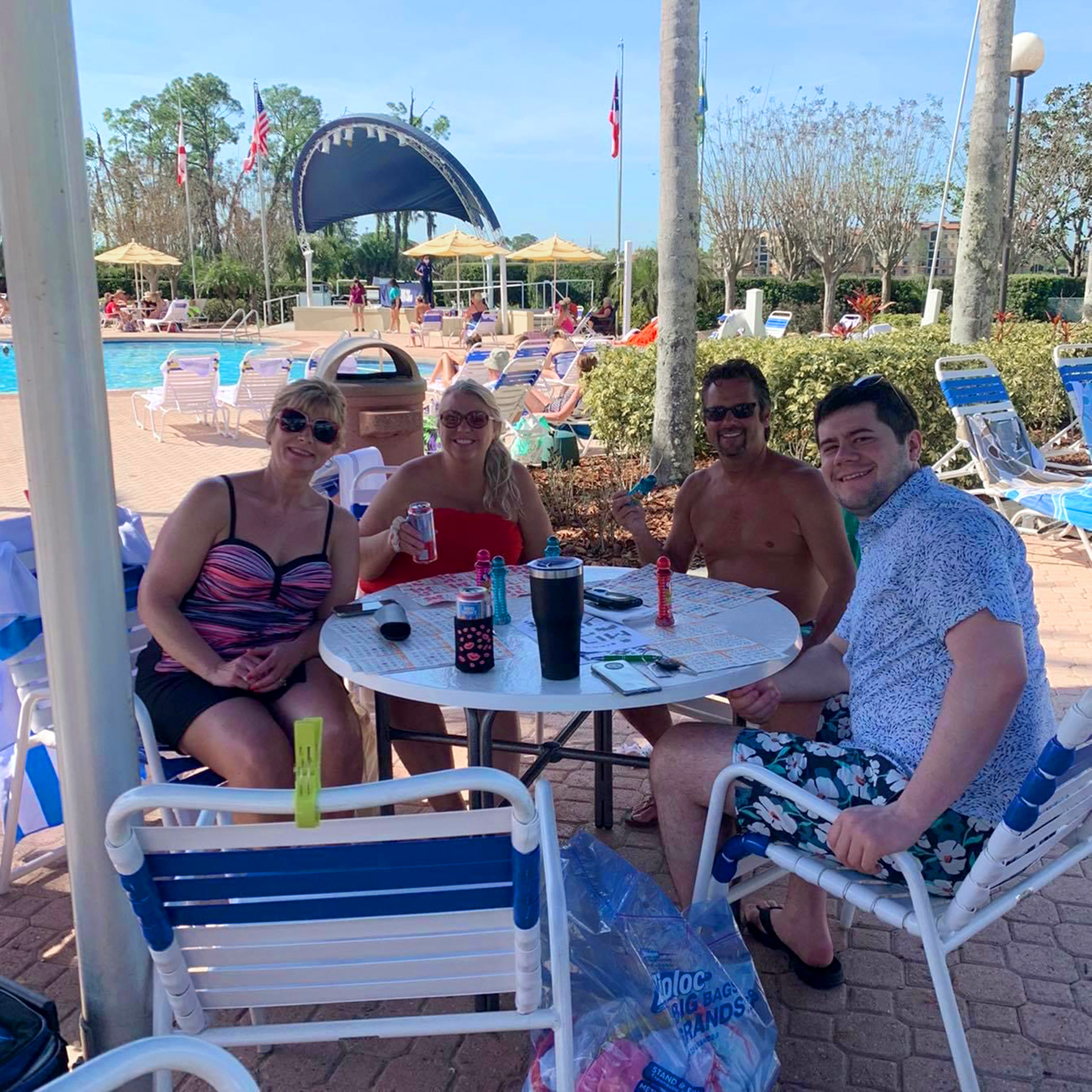 Two caucasian women wearing various colored swimsuits (left) and two caucasian men wearing trunks and a button-up (right) are seated around a poolside table underneath an umbrella with beverages.