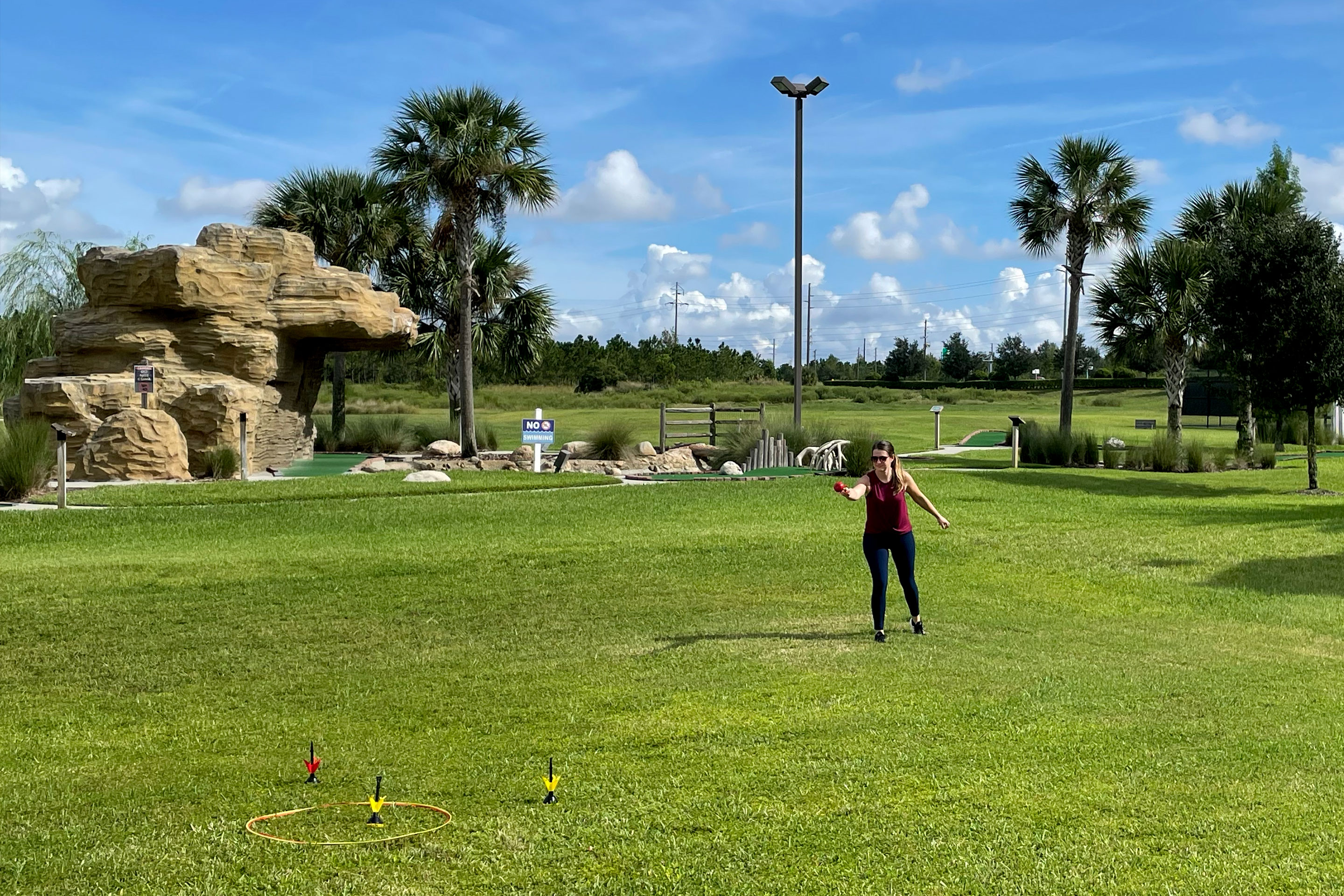 a woman in a red tank top and black leggings throws lawn darts while standing in an open grass field.