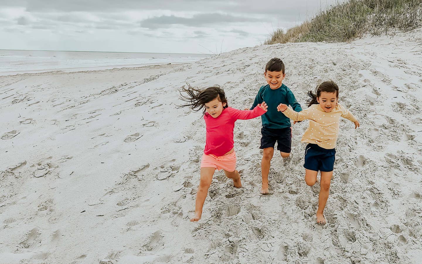 Three toddlers in multi-colored rash guards and board shorts run on a sandy beach.