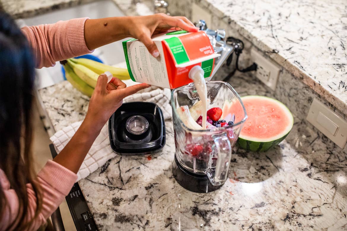 A woman fills a blender with ingredients for a smoothie at a kitchen counter.